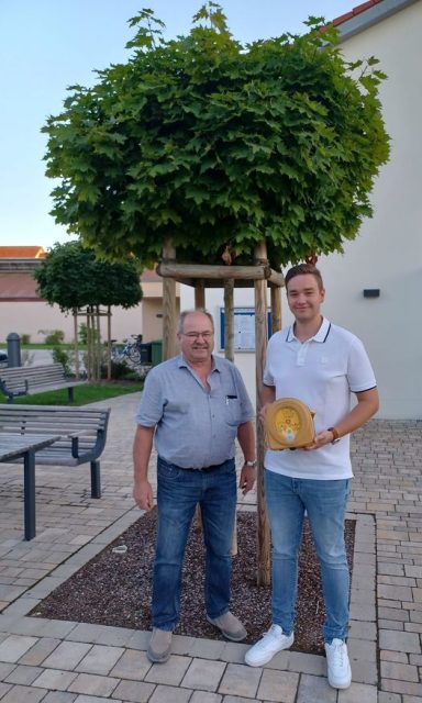 Zwei Männer stehen neben einem kleinen Baum, einer hält eine Auszeichnung in der Hand.